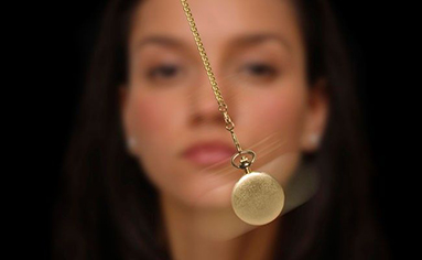 Mulher olhando fixamente para um pêndulo dourado em movimento, representando um tipo de sessão de hipnose.